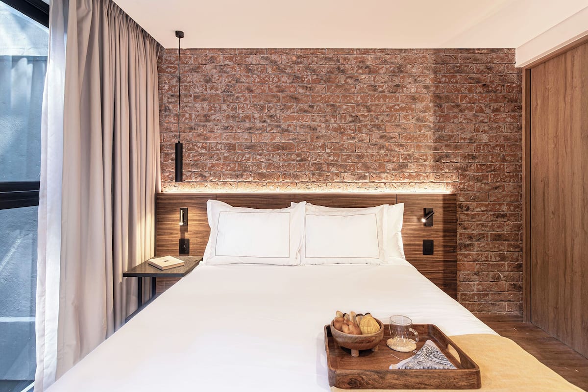 A hotel room with a brick wall, a bed with white linens, a nightstand with a lamp, and a breakfast tray with bread and a glass.