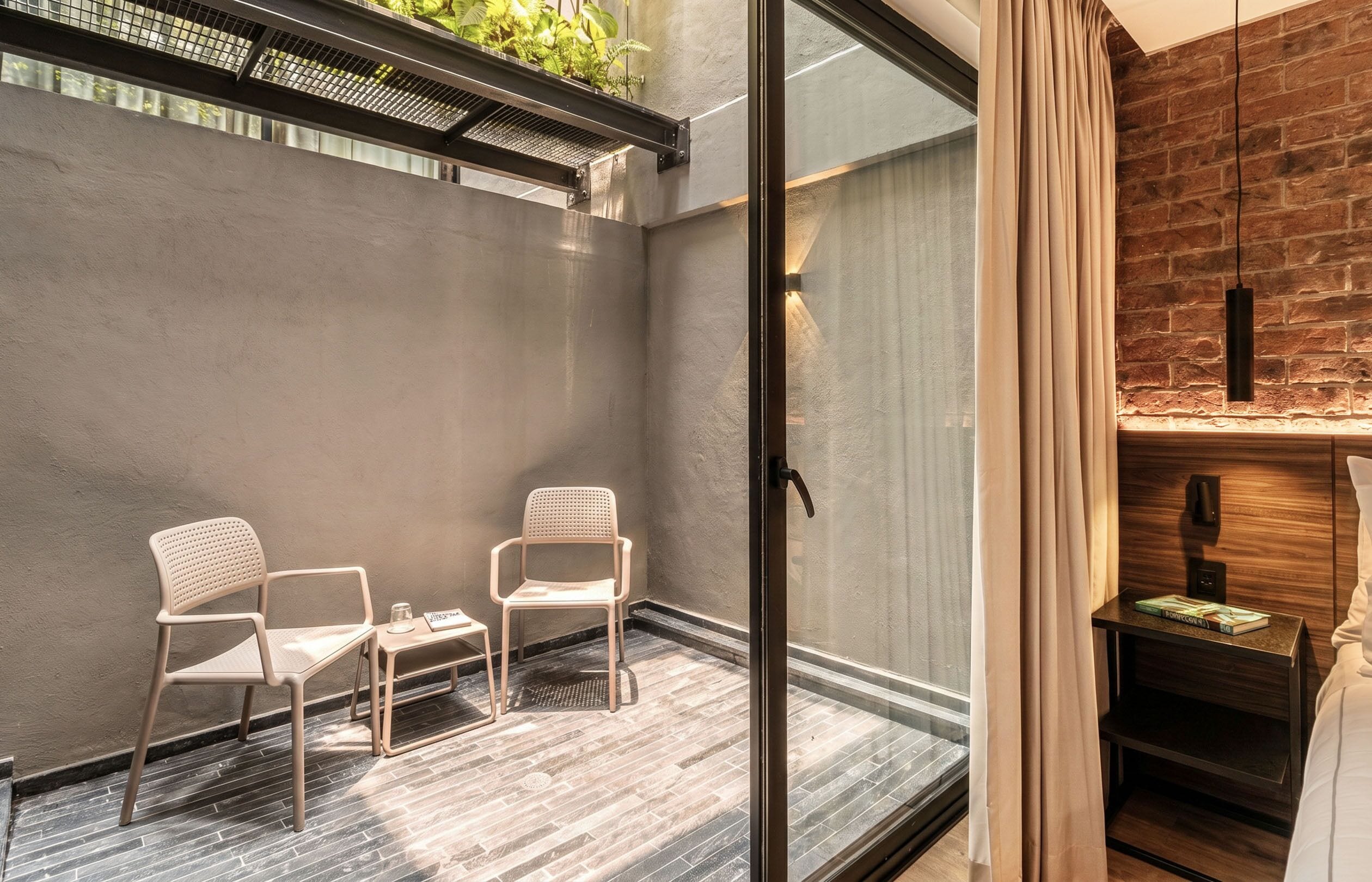 Una habitación moderna con puerta de cristal que da a un balcón, dos sillas, una mesita pequeña y una mesita de noche de madera con libros.