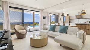 Una sala de estar moderna con sofá, mesa de centro y una silla, además de una zona de cocina y grandes ventanales con vista al océano.