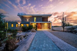 A modern two-story house with a stone facade, a paved driveway, and a desert landscape with cacti. - Villa Marah - Luxury Villa with Rooftop Terrace & Ocean Views (Kralendijk)