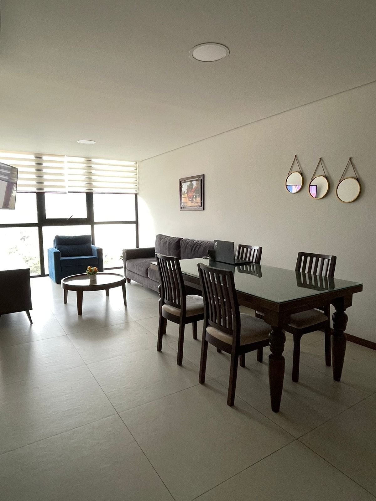 A living room with a sofa, a coffee table, a dining table with chairs, a television, and a window with blinds.