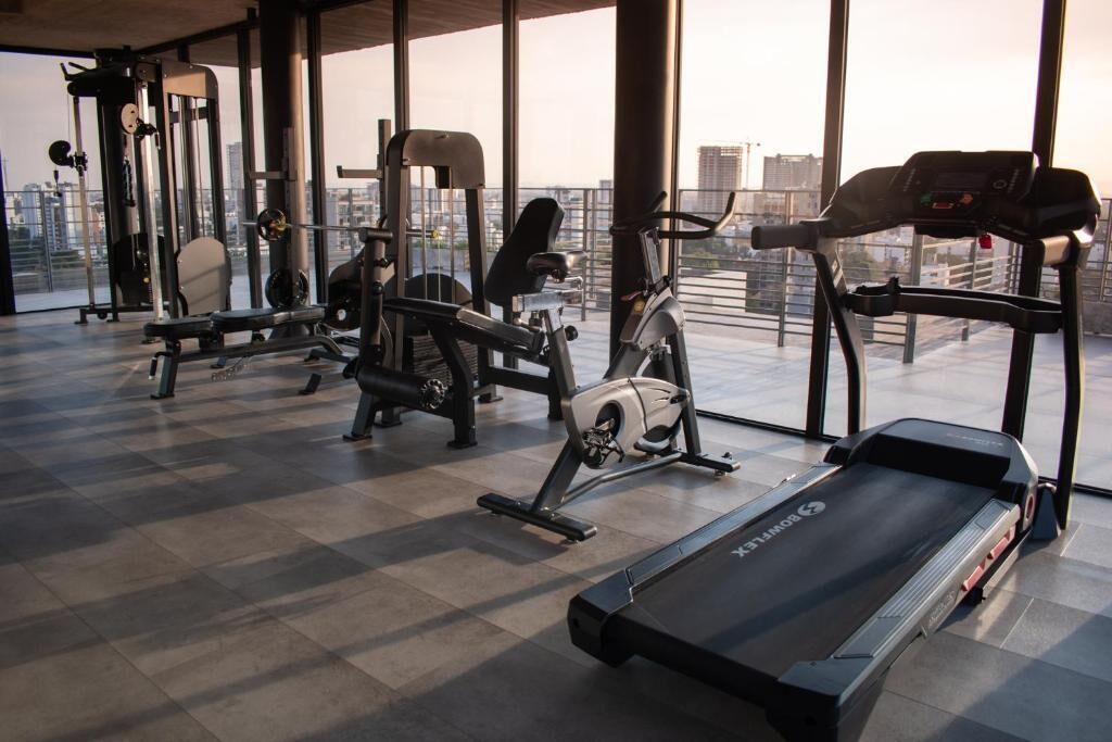 A gym with various exercise equipment, including a treadmill, stationary bikes, and weight machines, with a city view through large windows.