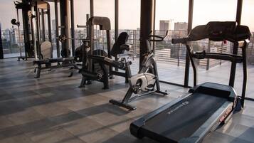 A gym with various exercise equipment, including a treadmill, stationary bikes, and weight machines, with a city view through large windows.