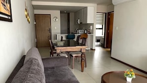 Una sala moderna con un sofá gris, una mesa de comedor con sillas, una cocina al fondo y una mesa redonda de centro con flores.