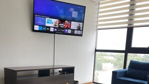 Una sala de estar moderna con un televisor de pantalla plana montado en la pared, un mueble de entretenimiento de madera oscura, una mesa de centro redonda de vidrio con un jarrón de flores, un sofá gris y un sillón azul.