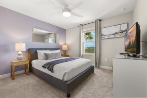 Une chambre à coucher comprenant un lit, des lampes de chevet, un ventilateur de plafond, un téléviseur, une fenêtre avec des rideaux et des œuvres d’art accrochées au mur.