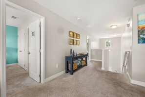 A hallway with carpeted floors, white doors, a blue console table with baskets, and framed pictures on the wall. - The Ultimate Private Oasis & Game Room 6 BD/ 3.5 BA/ Solterra Resort (5140 OA) (Davenport)