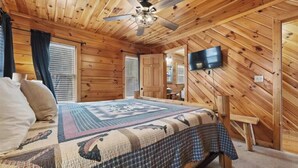 Een slaapkamer in een houten chalet, met een bed, een plafondventilator, een televisie en een raam met zonwering.