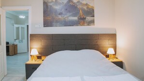 Habitación de hotel con cama, dos lámparas de noche, un cuadro de un velero en el agua y vista a un pasillo.