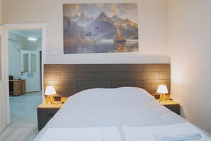 Une chambre d’hôtel avec un lit, deux lampes de chevet, un tableau représentant un voilier sur l’eau et une vue sur un couloir.