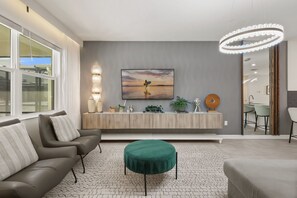 A modern living room with a sofa, a round ottoman, a dining area with chairs, a kitchen visible through an open door, and a large painting on the wall. - Custom Theme Rooms 9 BD/5 BA/Storey Lake - 4520 TB (Kissimmee)