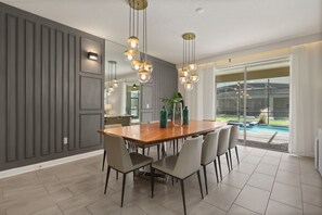 A modern dining area with a wooden table, six chairs, pendant lighting, and a view of a swimming pool. - Custom Theme Rooms 9 BD/5 BA/Storey Lake - 4520 TB (Kissimmee)