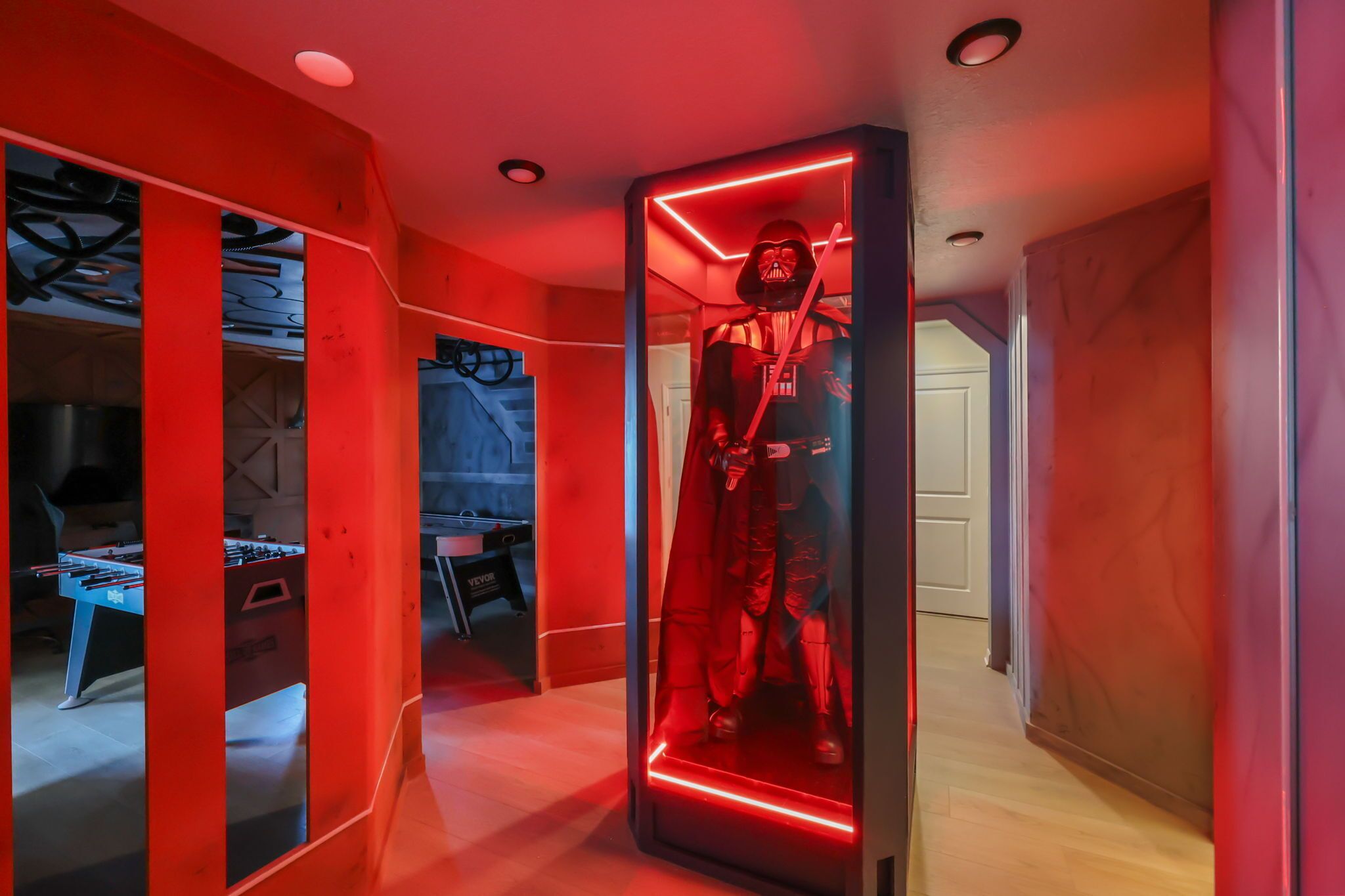 Una habitación con un expositor iluminado en rojo que contiene una figura de Darth Vader, una mesa de futbolín y una zona de cocina visible.