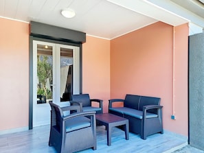 Une terrasse couverte avec des meubles en osier, une porte-fenêtre coulissante donnant sur un jardin et un luminaire au plafond.