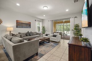 A modern living room with a sofa, armchairs, a coffee table, and a TV. There is a painting on the wall and a view of the outdoors through large windows. - Private Pool! 7 BD/ 5.5 BA/ Windsor at Westside (1913 NC) (Kissimmee)