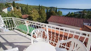 A balcony with white railings, a green area, and a view of a lake and buildings. - Cozy apartment in Omisalj with WiFi (Omisalj)
