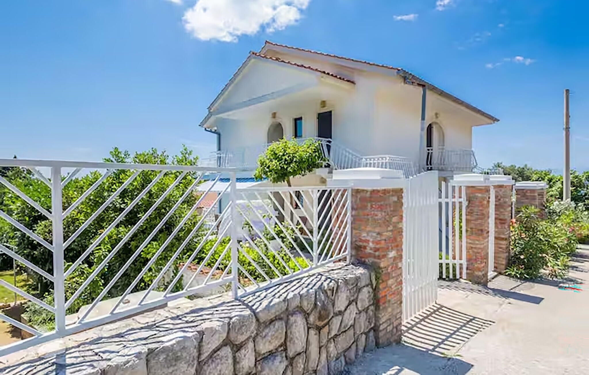 Une villa blanche avec une entrée surmontée d’un mur de pierre, un balcon et un ciel d’un bleu limpide.