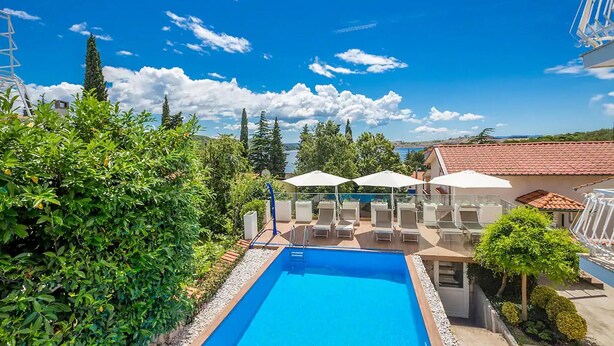 A swimming pool with lounge chairs and umbrellas, surrounded by greenery and overlooking a body of water. - Cozy apartment in Omisalj with WiFi (Omisalj)