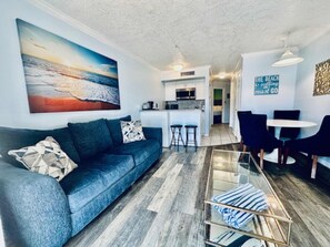 A living room with a sofa, a glass coffee table, a painting, a kitchen area, and a dining table with chairs. - Sandy Seawall Escape Resort (Galveston)