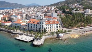 A coastal cityscape with a prominent hotel featuring a red-tiled roof, a waterfront terrace, and a view of the sea.