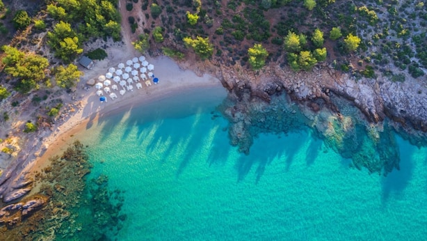 Une vue aérienne d’une plage aux eaux turquoise et limpides, avec une côte rocheuse et une rangée de parasols blancs.