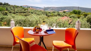 Une terrasse avec deux chaises orange, une petite table ronde sur laquelle se trouve un panier de fruits, et une vue sur un paysage vallonné.