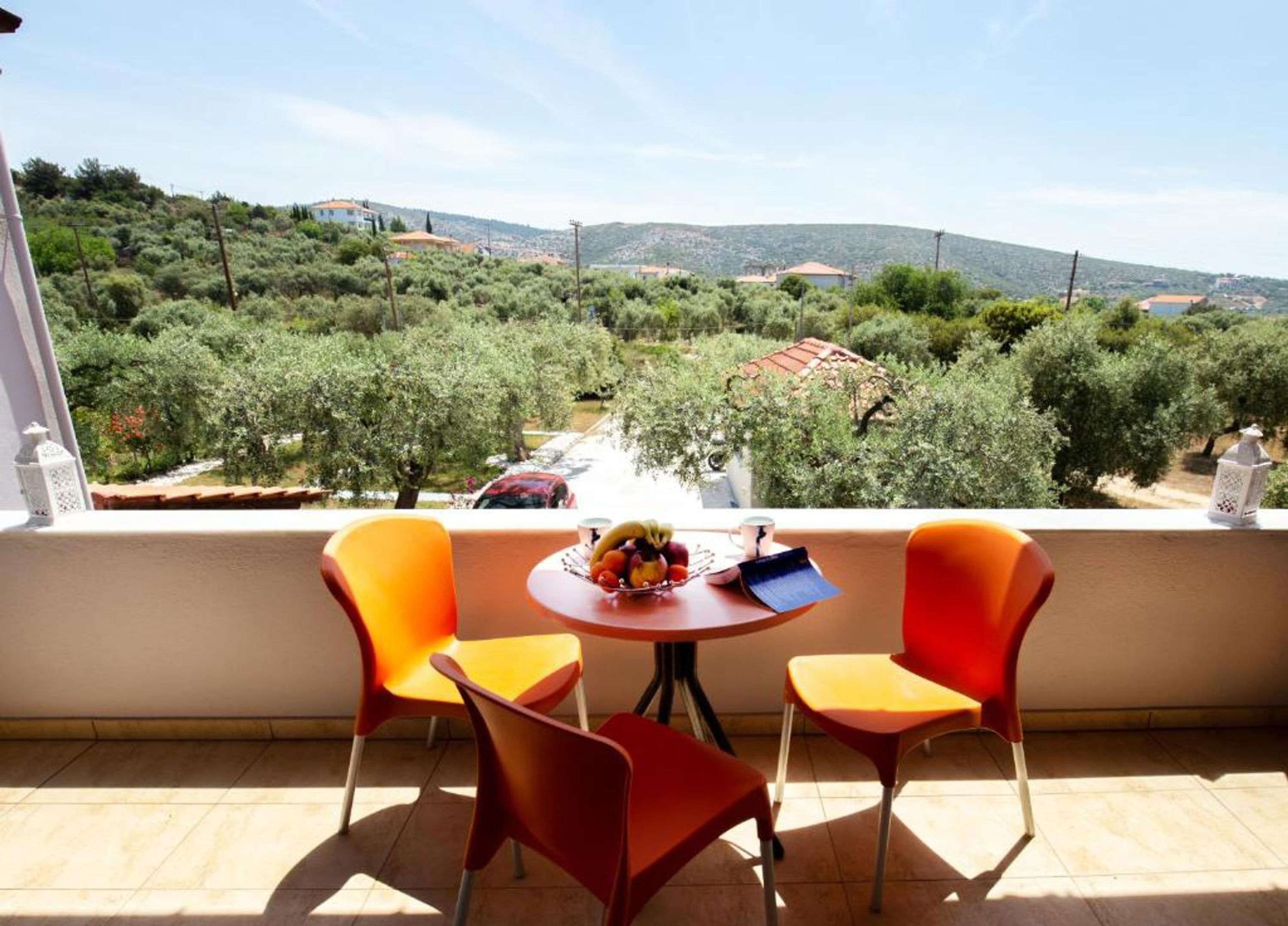A balcony with two orange chairs and a small table, overlooking a scenic landscape with trees and buildings.