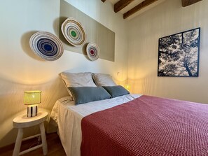 A bedroom with a bed, pillows, a red blanket, a small stool with a lamp, and wall decor including circular wall hangings and a framed picture.