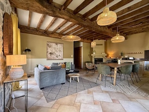 A rustic interior with exposed wooden beams, a dining area with a wooden table and chairs, a sofa, and a fireplace.