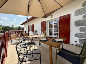 A wooden table with chairs and a large umbrella, set against a white wall with red shutters and stone accents.