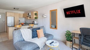 Ett modernt vardagsrum med en grå soffa, ett litet runt soffbord, en tv som visar ”NETFLIX”, ett köksområde med träskåp och en mikrovågsugn.