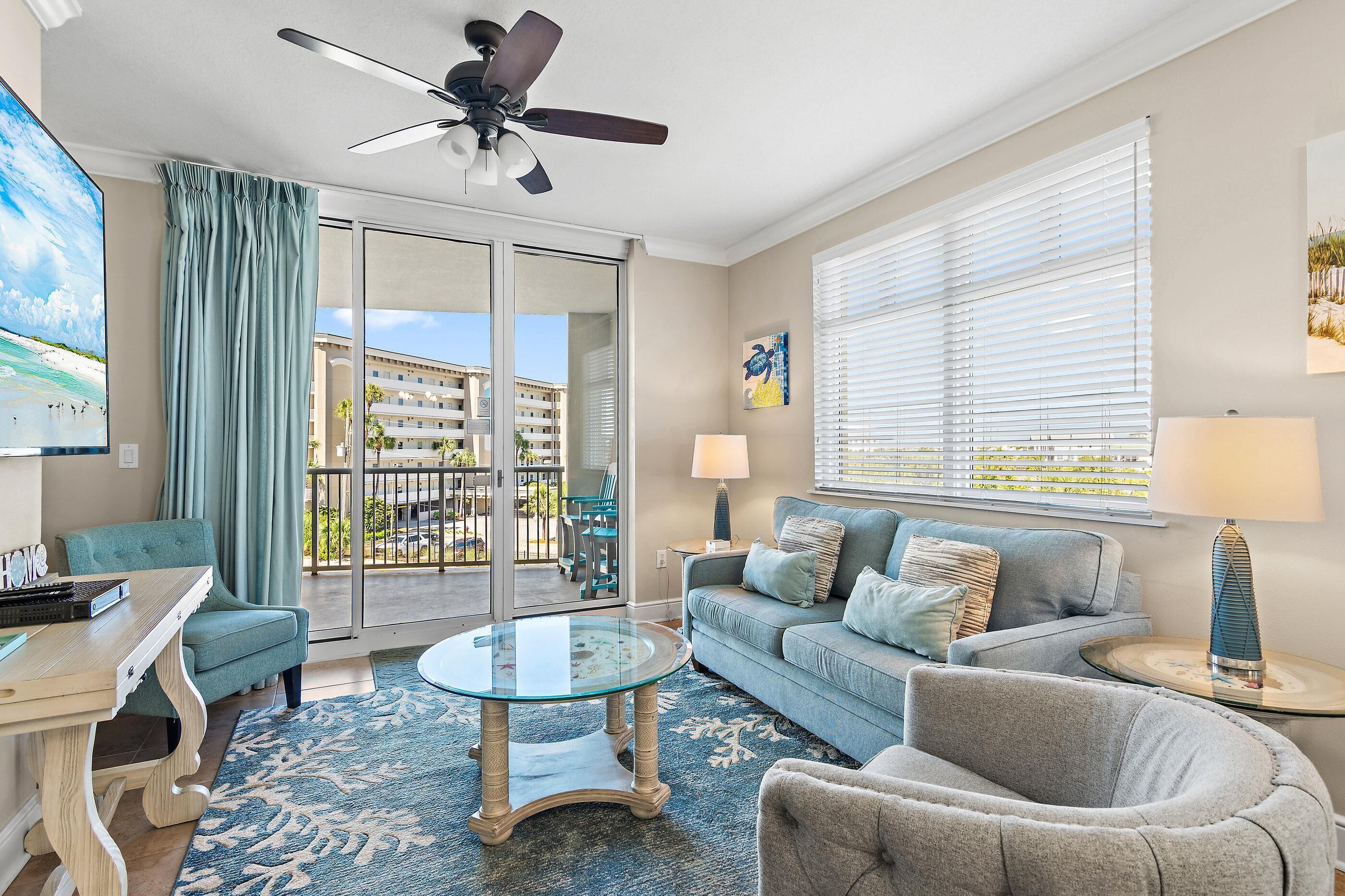 A living room with a glass coffee table, a sofa, a chair, a ceiling fan, a TV, and a balcony with a view of a building.
