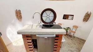Eine kompakte Küche mit eingebautem Herd, Spüle und Kühlschrank, ein Fenster mit blickdichter Scheibe und eine kleine Sitzecke mit Stuhl und Tisch.
