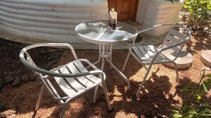 Ein kleiner Außenbereich mit Sitzgelegenheit, einem runden Tisch, zwei Metallstühlen, einem Blumentopf und einem Gebäude mit einer Holztür im Hintergrund.