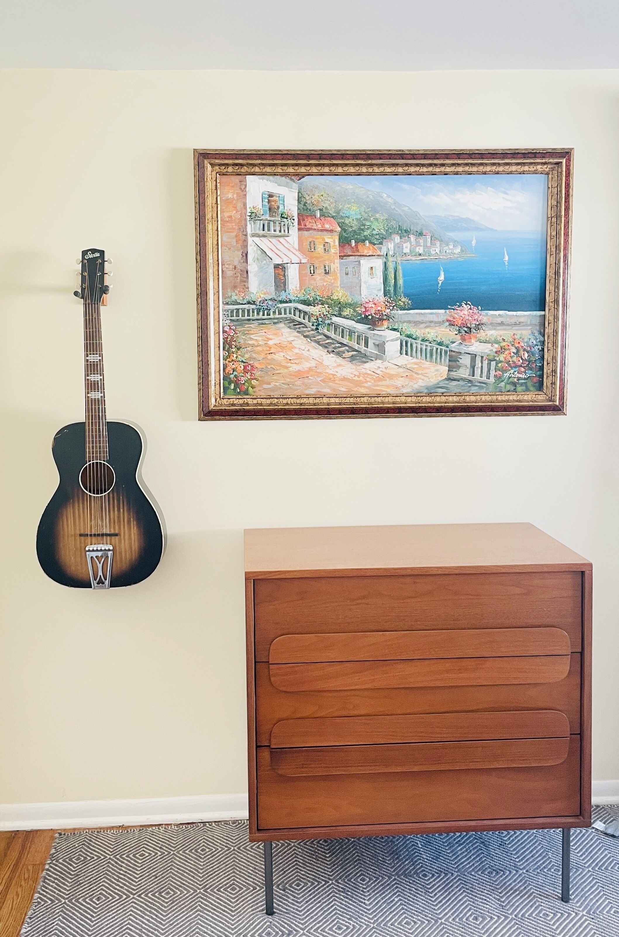 Een gitaar, een lijst met een schilderij en een houten kast tegen een muur.