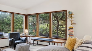 A room with large windows, a black leather chair, a wooden bench, and a bookshelf with books. - Magic Tree House by Avantstay Fire Pit + Deck + Movie Room Walk to the Beach (Manzanita)