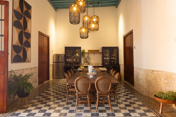 A dining area with a checkered floor, a long table with chairs, hanging light fixtures, and wooden cabinets.