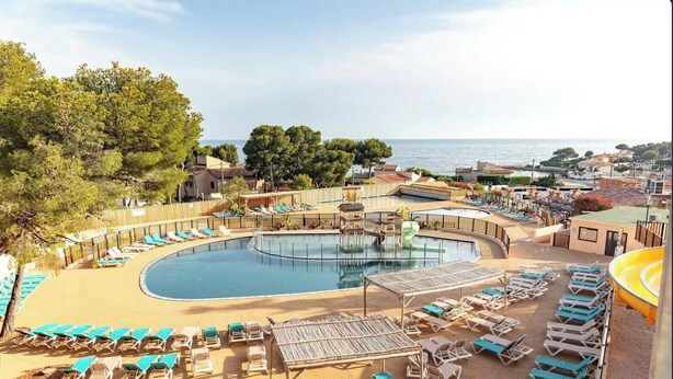 Uma área de piscina com espreguiçadeiras e uma piscina circular, cercada por edifícios e árvores.
