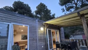 Um deck de madeira com mesa e cadeiras, uma porta de vidro deslizante que dá para um espaço interno e uma área coberta com um banco.