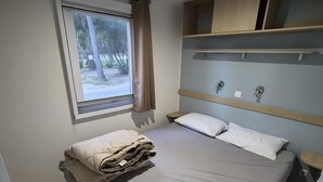 Uma cama de solteiro com um cobertor bem dobrado, uma janela com cortinas, uma prateleira embutida e um aparelho de luz fixado na parede.