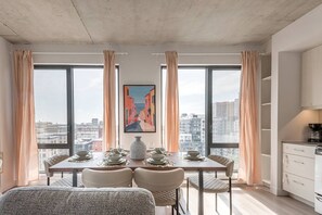 A dining area with a table set for four, large windows with curtains, a painting on the wall, and a view of buildings outside. - Skyline Penthouse | 360° Views (Montreal)