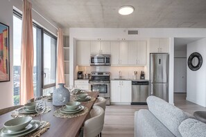 A modern kitchen with white cabinets, stainless steel appliances, and a dining area with a wooden table set for four. - Skyline Penthouse | 360° Views (Montreal)