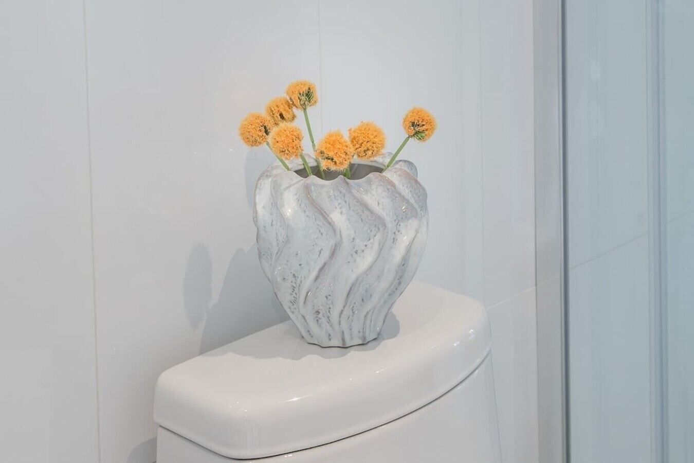 A white ceramic vase with a wavy design containing yellow flowers on a white toilet.