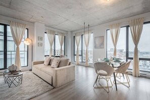 A modern living room with a sofa, dining table, chairs, and large windows with curtains. - Skyline Penthouse | 360° Views (Montreal)