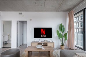 A modern living room with a flat-screen TV displaying the Netflix logo, a wooden coffee table, a potted plant, and a large window with curtains. - Skyline Penthouse | 360° Views (Montreal)