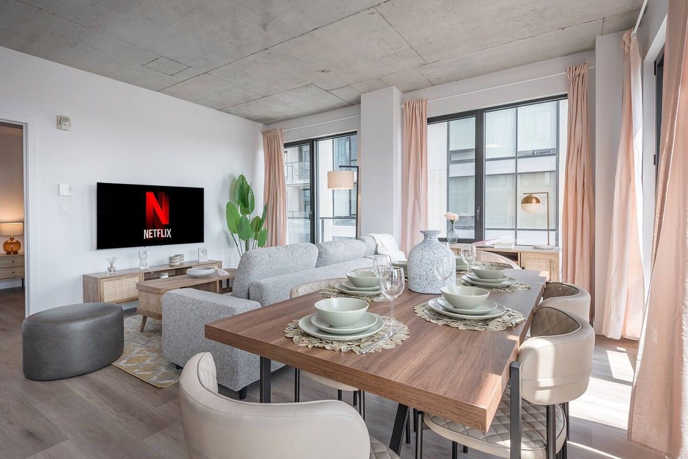 A modern living room with a flat-screen TV, a sofa, a dining table set for four, and a large window with curtains.