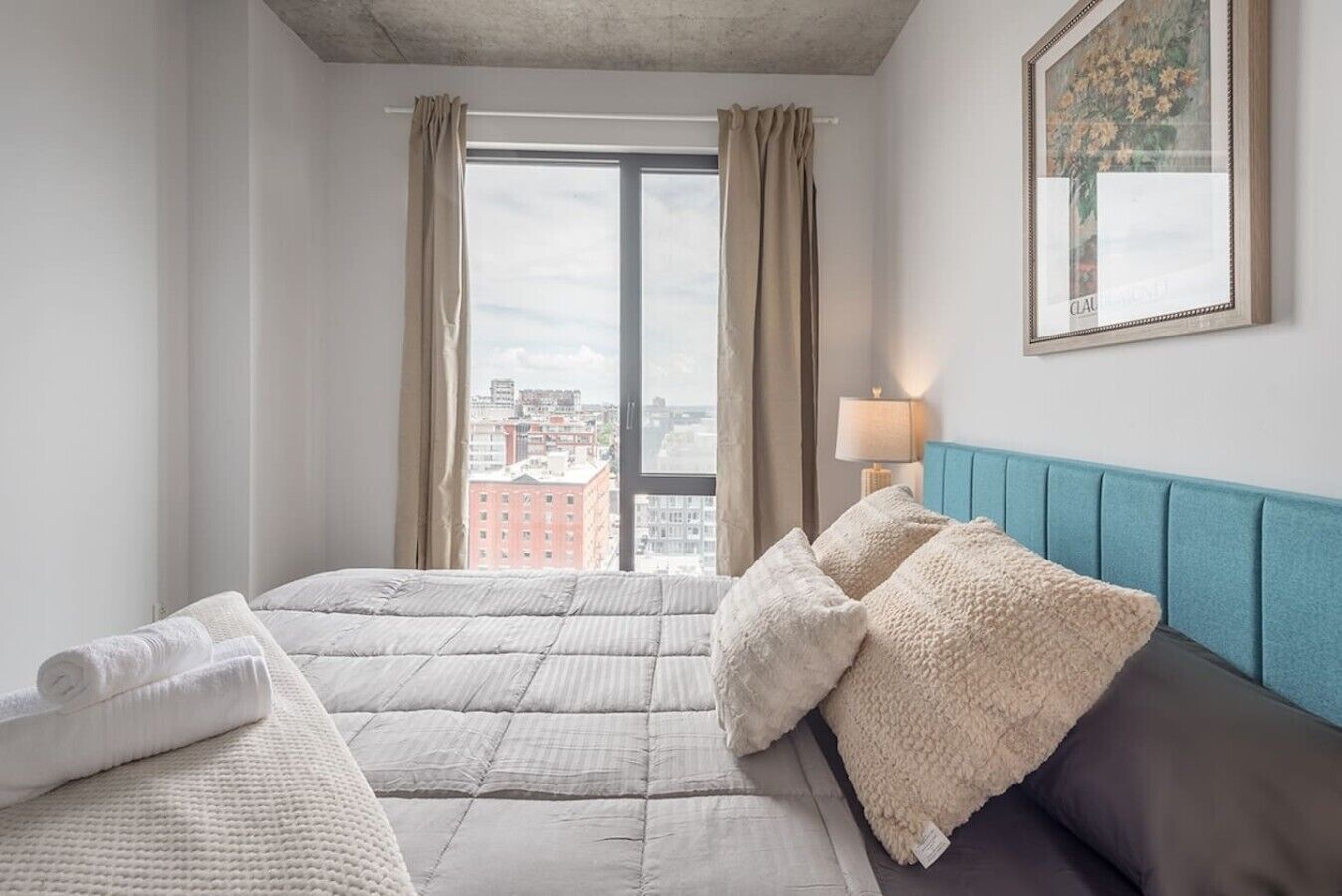 Et hotelværelse med en seng, et blåt sengegavl, et vindue med gardiner, en lampe og et billede i ramme på væggen.