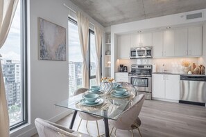 A modern kitchen with stainless steel appliances, a dining area with a glass table, and a city view. - Skyline Penthouse | 360° Views (Montreal)