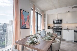 A modern kitchen with white cabinets, stainless steel appliances, and a dining area with a table set for four. - Skyline Penthouse | 360° Views (Montreal)