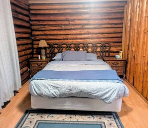 Une chambre dans un chalet en rondins, avec une tête de lit en bois, un lit recouvert d’une literie à motifs floraux, deux tables de chevet avec des lampes et un tapis à motifs.